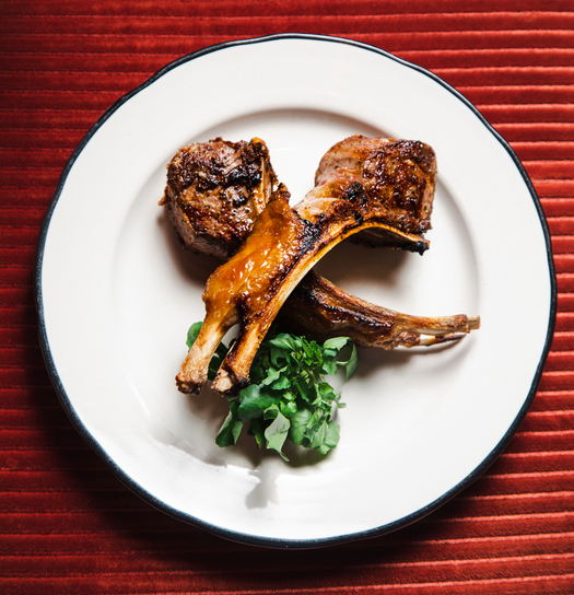 Grilled lamb chops with fresh greens on a white plate with black rim, set on red textured tablecloth.