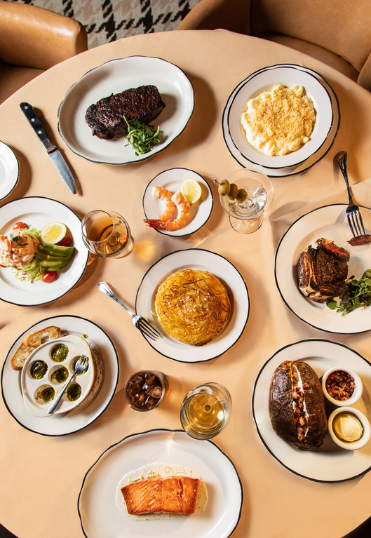 A variety of dishes including steak, shrimp, omelette, baked potatoes, and bread with snails on a round table.