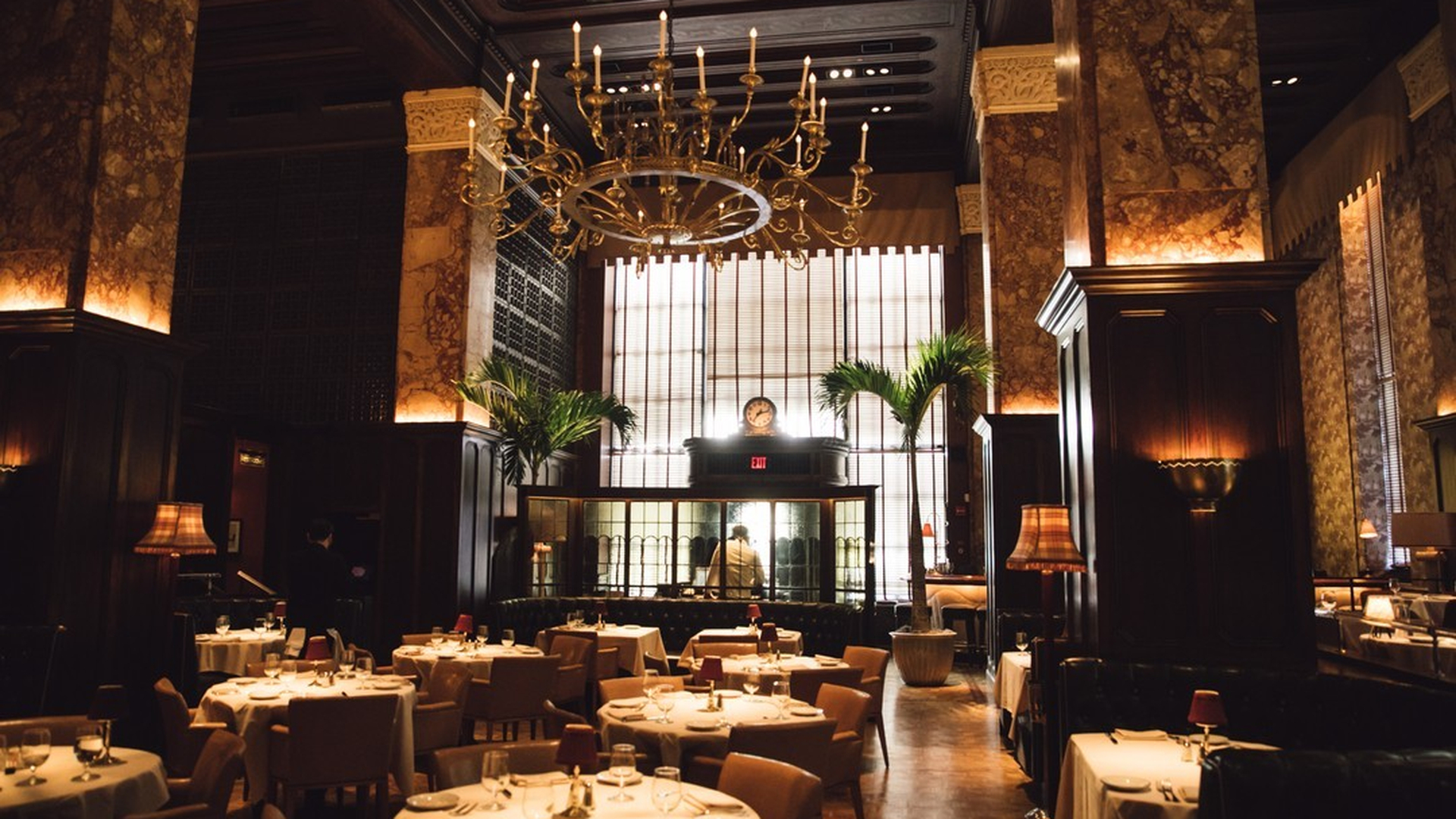 Elegant restaurant interior with marble walls, chandelier, large windows, and set tables for dining.