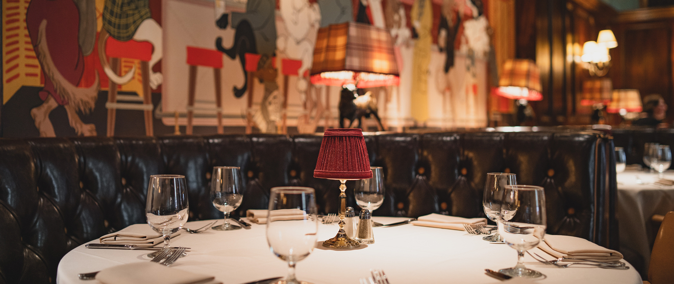 Elegant restaurant table set with white linens, glasses, and napkins against a backdrop of black leather booths and vibrant wall art.