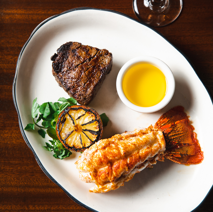 Grilled steak, lobster tail, grilled pineapple, garnished with herbs, lemon butter on a white plate.