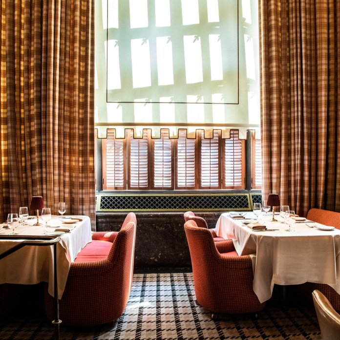 Chic restaurant interior with red chairs, white tablecloths, checkered floor, and patterned window, warm lighting.